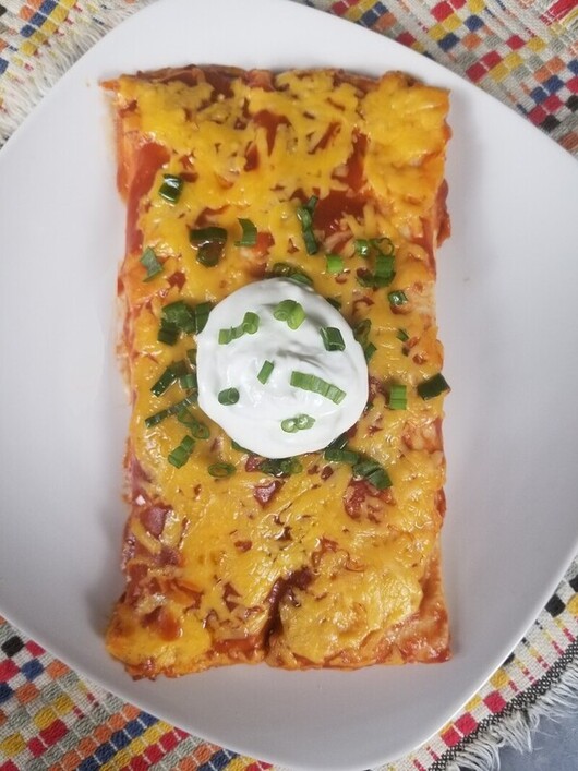 two cheesy chicken enchiladas with red sauce on a white plate with sour cream and green onions
