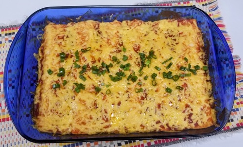 six baked chicken enchiladas rolled up in a baking dish covered with red sauce and shredded cheddar cheese, topped with chopped green onions
