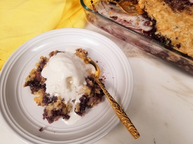 blackberry cobbler dump cake on a plate with vanilla ice cream next to a cake pan of blackberry cobbler