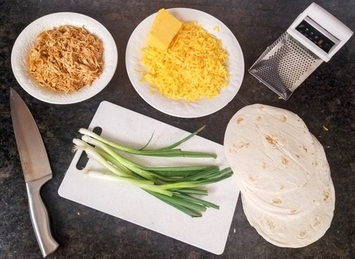 cheesy chicken enchilada ingredients, a bowl of shredded cheddar cheese, a bowl of shredded chicken, flour tortilla shells, green onions with a cutting board and knife and cheese shredder all on a black counter top 