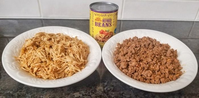 a bowl of shredded chicken, a bowl of ground beef and a can of refried beans on a black counter top