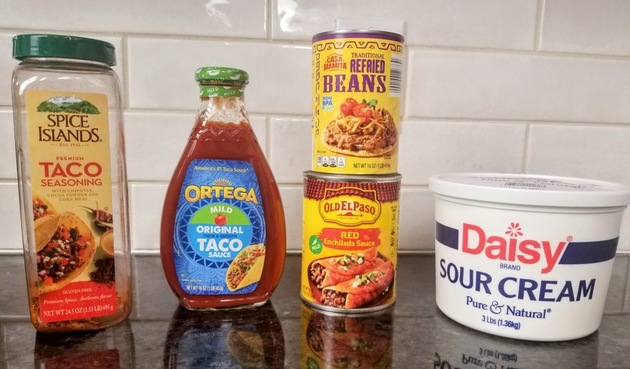 taco seasoning, a bottle of taco sauce, sour cream, a can of red enchilada sauce, and a can of refried beans on a black counter top