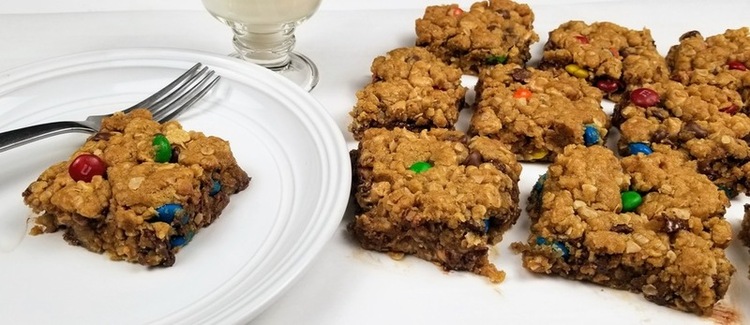 a monster cookie bar on a white plate surrounded by other bars and a glass of milk on a white counter
