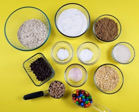 flour, white and brown sugar, baking soda, baking powder, rice krispies, oatmeal, chocolate chips, peanut butter chips and M&M's in seperate bowls on a yellow counter