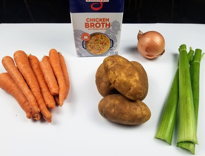 carrots, potatos, celery, an onion, and a carton of chicken broth on a white counter
